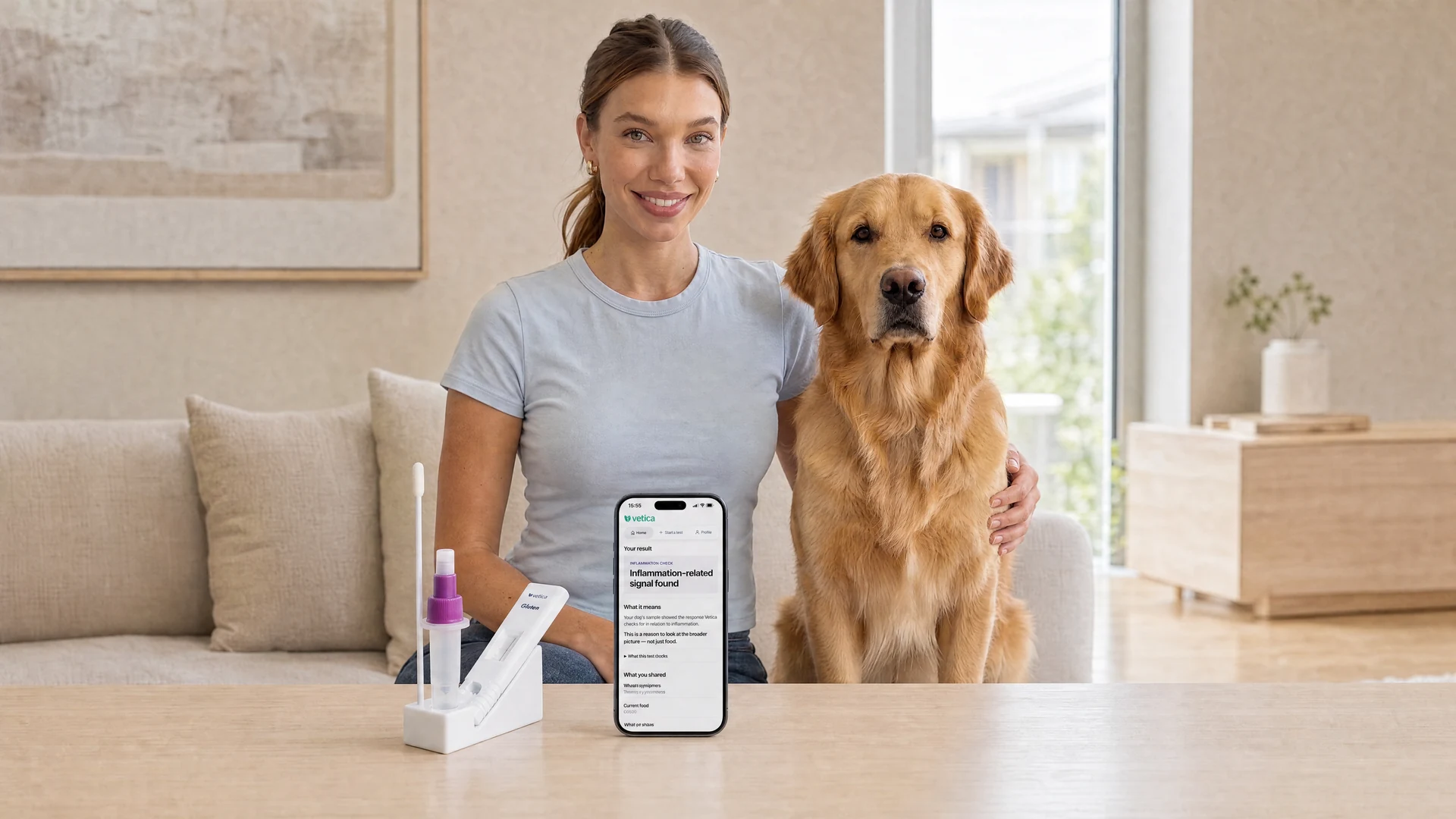 A woman and her golden retriever facing the camera at home, with a Vetica saliva test kit and a phone displaying a Vetica report on the table